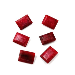 May include: Six red rectangular gemstones, likely rubies, are arranged on a white background. The gemstones are all the same size and shape, and they are all cut in a rectangular emerald cut.