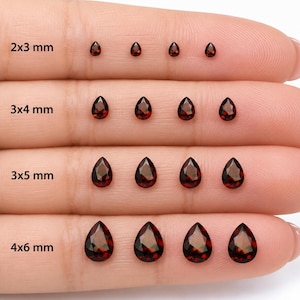 May include: A collection of sixteen deep red, pear-shaped faceted gemstones arranged in four rows by size. The rows are labeled 2x3 mm, 3x4 mm, 3x5 mm, and 4x6 mm, indicating the specific metric dimensions for each set of four identical stones.