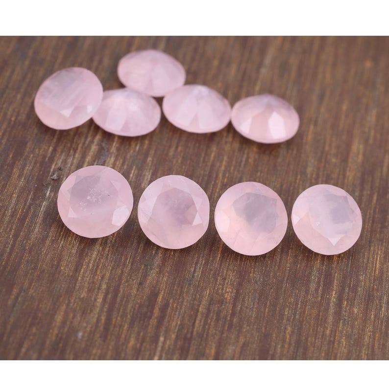 Puede incluir: Un conjunto de gemas de cuarzo rosa claro facetadas. Las gemas son ovaladas y redondas, con una superficie pulida y reflectante. Las piedras est&aacute;n dispuestas sobre una superficie de madera marr&oacute;n oscuro.