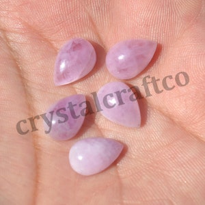May include: Five teardrop-shaped pink gemstone cabochons, each with a smooth, polished surface. The stones are arranged in a hand, with the text "crystalcraftco" visible in the background.