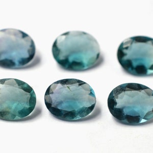 May include: Six oval-cut gemstones with a teal and blue color gradient. The gems are arranged on a white surface, showcasing their polished facets and translucent appearance. These gemstones are suitable for jewelry making.