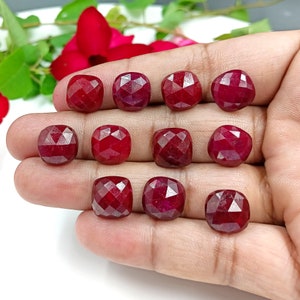 May include: Ten faceted red ruby gemstones, each with a square shape, are displayed on a hand. The gemstones are arranged in two rows of five.
