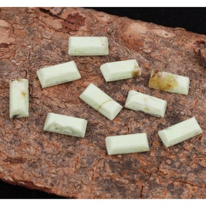 May include: Ten light green rectangular cabochon gemstones on a brown wooden surface.