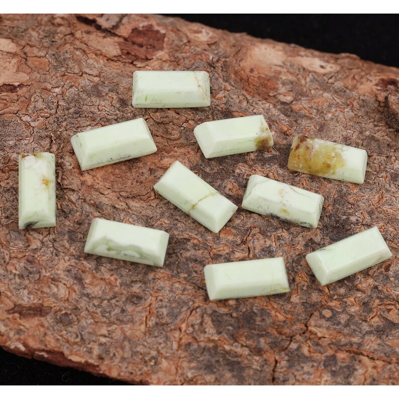 May include: Ten light green rectangular cabochon gemstones on a brown wooden surface.