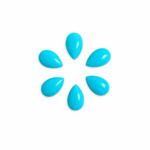 Puede incluir: Cinco cabujones de color turquesa brillante, lisos y con forma de lágrima, están dispuestos en un patrón circular. Cada pieza presenta una superficie brillante y reflectante con una parte superior redondeada y una base puntiaguda, creando un conjunto uniforme de piedras azul verdoso.