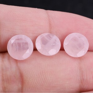Puede incluir: Tres gemas redondas facetadas de cuarzo rosa. Las piedras son de color rosa suave y tienen una superficie pulida y reflectante. Las gemas se muestran sobre un fondo neutro.