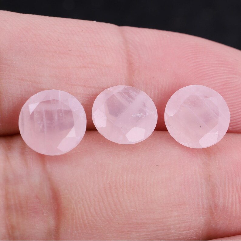 Puede incluir: Tres gemas redondas facetadas de cuarzo rosa. Las piedras son de color rosa suave y tienen una superficie pulida y reflectante. Las gemas se muestran sobre un fondo neutro.
