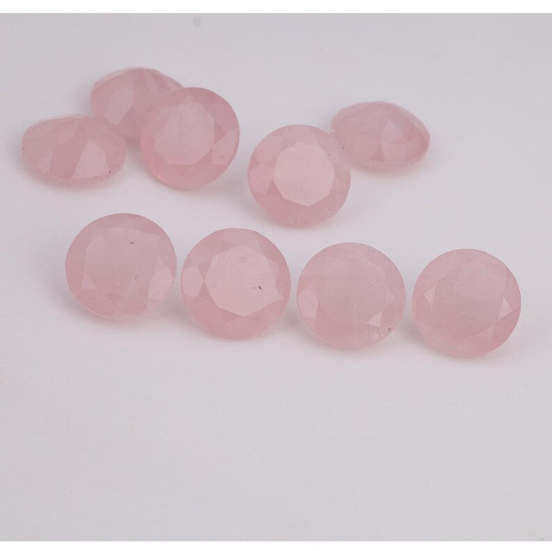 Puede incluir: Una colecci&oacute;n de gemas redondas de cuarzo rosa facetadas. Las gemas son de color rosa suave y tienen una superficie pulida y reflectante. Las piedras est&aacute;n dispuestas sobre un fondo blanco.
