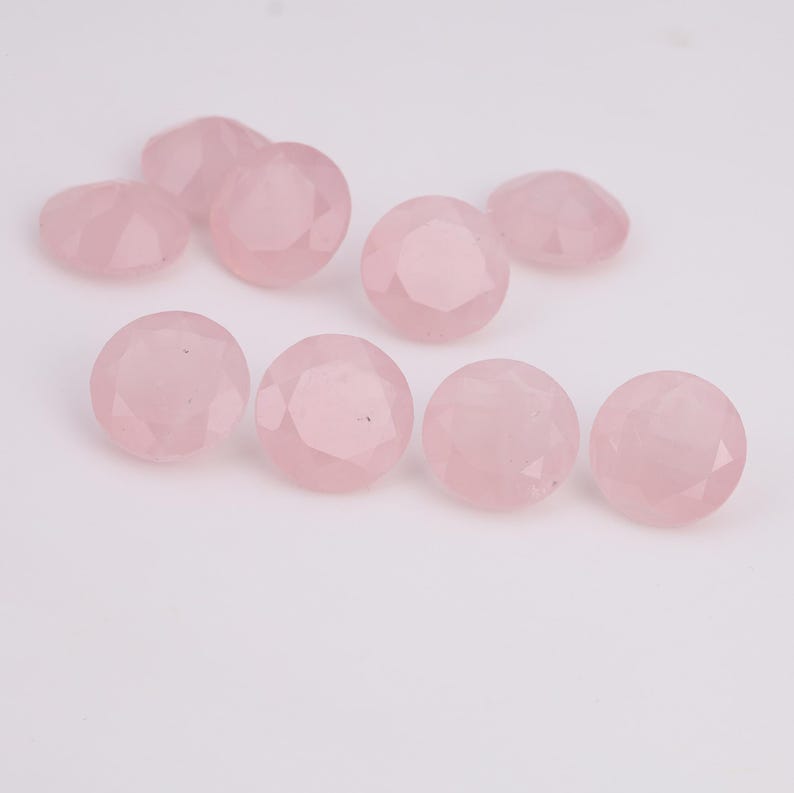 Puede incluir: Una colecci&oacute;n de piedras preciosas de cuarzo rosa facetadas de color rosa claro. Las piedras preciosas son redondas y tienen m&uacute;ltiples facetas que capturan la luz. Las piedras est&aacute;n dispersas sobre una superficie blanca.
