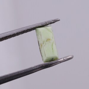 May include: A light green gemstone with a rectangular shape, held by tweezers.