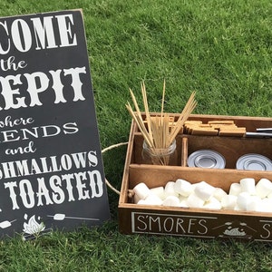 May include: A wooden box with the words "S'mores Station" on the front. The box is filled with marshmallows, graham crackers, and chocolate bars. There is a sign that says "Welcome to the Firepit where friends and marshmallows get toasted".