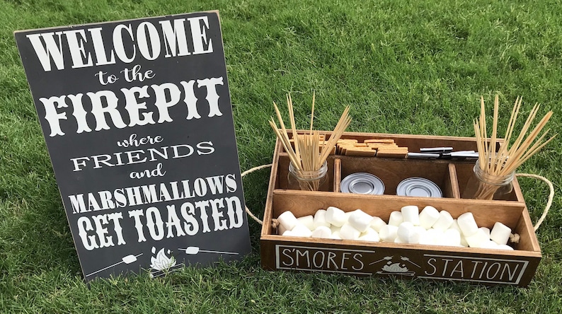 May include: A wooden box with the words "S'mores Station" on the front. The box is filled with marshmallows, graham crackers, and chocolate bars. There is a sign that says "Welcome to the Firepit where friends and marshmallows get toasted".