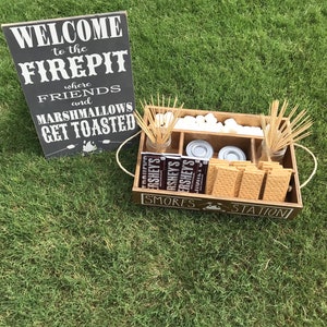 May include: A wooden tray with the words "S'mores Station" on the front. The tray is filled with graham crackers, marshmallows, chocolate bars, and skewers. A sign behind the tray reads "Welcome to the Firepit where friends and marshmallows get toasted."