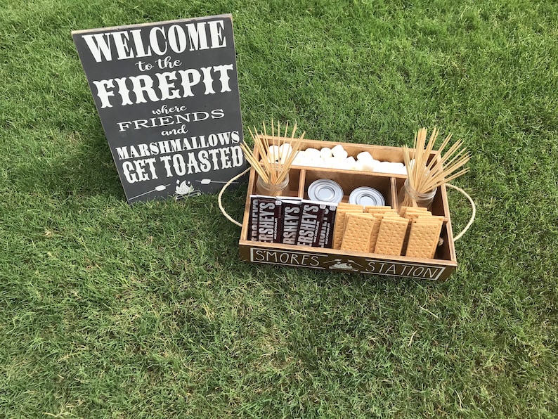 May include: A wooden tray with the words "S'mores Station" on the front. The tray is filled with graham crackers, marshmallows, chocolate bars, and skewers. A sign behind the tray reads "Welcome to the Firepit where friends and marshmallows get toasted."