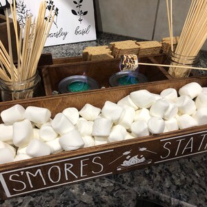 May include: A wooden box with the words "S'mores Station" on the front. The box is divided into sections and contains marshmallows, graham crackers, chocolate bars, and skewers.