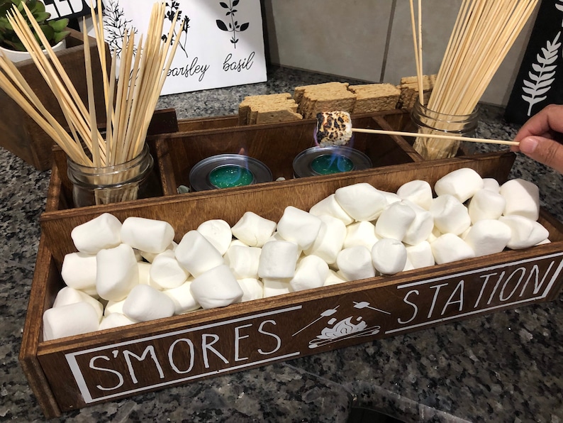 May include: A wooden box with the words "S'mores Station" on the front. The box is divided into sections and contains marshmallows, graham crackers, chocolate bars, and skewers.
