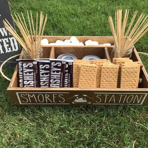 May include: A wooden tray with compartments for making s'mores. The tray has a sign that says "S'mores Station". The tray contains graham crackers, chocolate bars, marshmallows, and skewers.