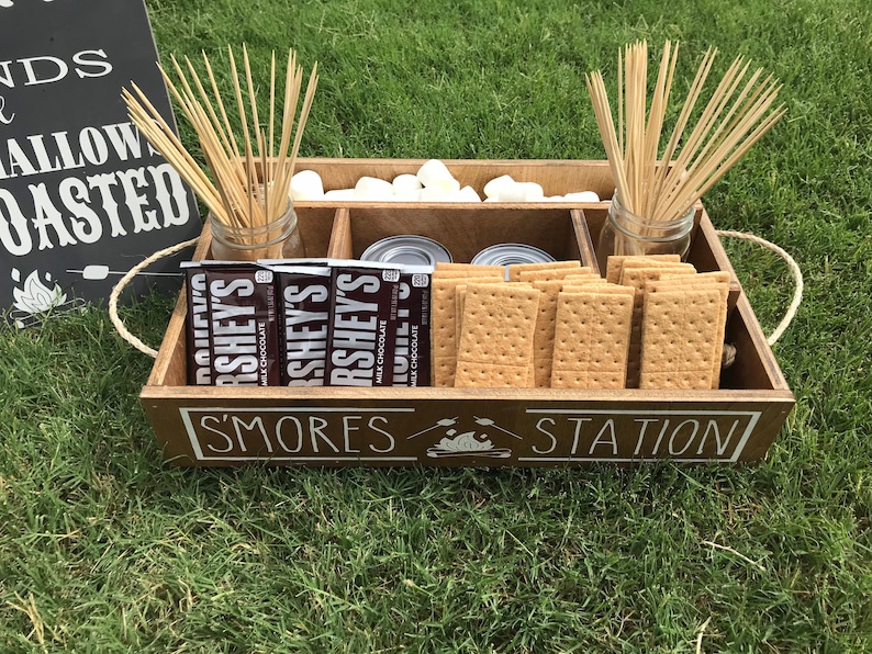 May include: A wooden tray with compartments for making s'mores. The tray has a sign that says "S'mores Station". The tray contains graham crackers, chocolate bars, marshmallows, and skewers.