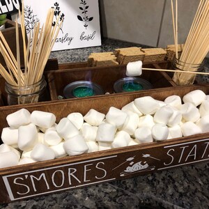 May include: A wooden box with the words "S'mores Station" on the front. The box is filled with white marshmallows, wooden skewers, and two small metal containers.