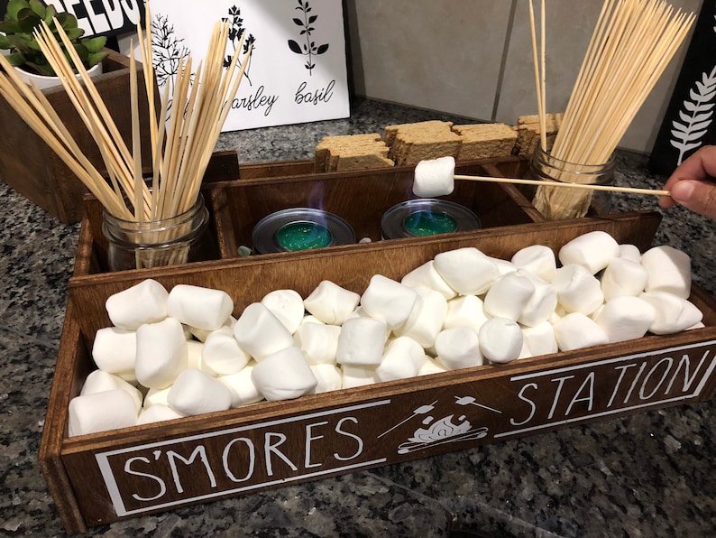 May include: A wooden box with the words "S'mores Station" on the front. The box is filled with white marshmallows, wooden skewers, and two small metal containers.