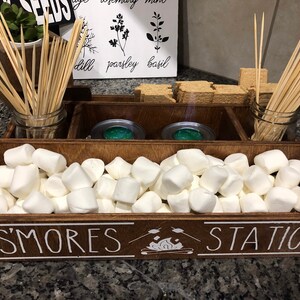 May include: A wooden box with the words "S'mores Station" on the front. The box is divided into sections and contains graham crackers, marshmallows, and skewers.