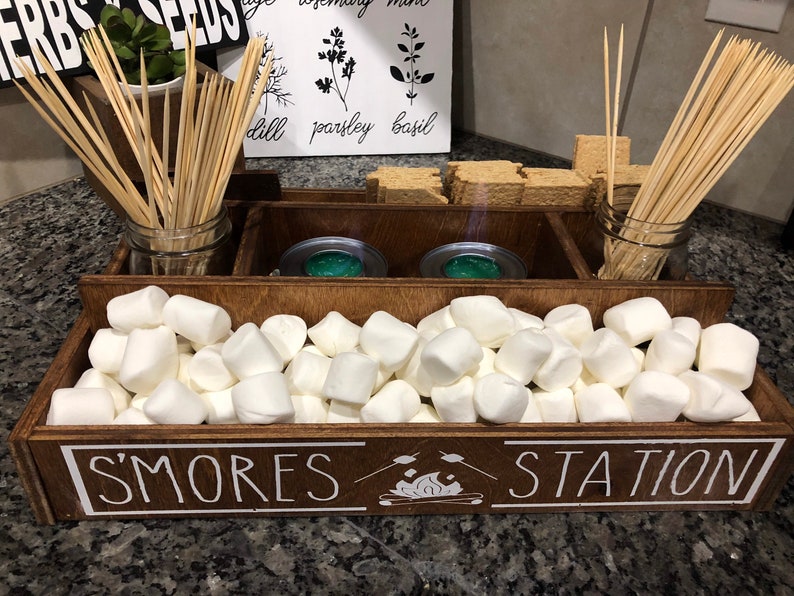 May include: A wooden box with the words "S'mores Station" on the front. The box is divided into sections and contains graham crackers, marshmallows, and skewers.