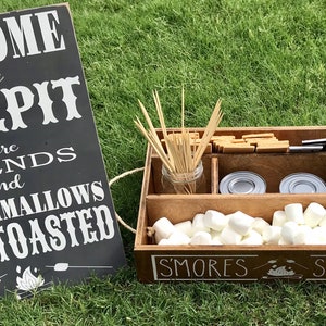 May include: A wooden s'mores station with a sign that reads "Welcome to the Firepit where friends and marshmallows get toasted." The station has compartments for marshmallows, graham crackers, chocolate, and skewers.