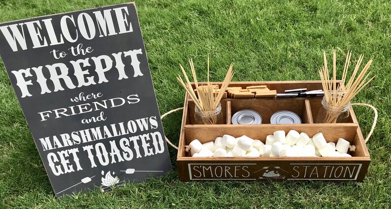 May include: A wooden s'mores station with a sign that reads "Welcome to the Firepit where friends and marshmallows get toasted." The station has compartments for marshmallows, graham crackers, chocolate, and skewers.