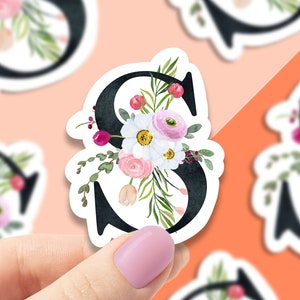 May include: A sticker featuring a black letter S with a watercolor floral design. The flowers are pink, white, and red. The sticker is on a white background.
