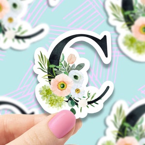 May include: A white sticker with a black letter C decorated with watercolor flowers in shades of pink, white, and green.