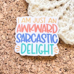 May include: A white sticker with the text "I am just an awkward sarcastic delight" in colorful lettering. The text is in a playful, handwritten font. The sticker is on a corkboard background.