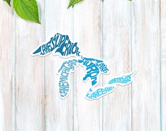 May include: A blue and white sticker of the Great Lakes with the names of each lake spelled out within the outline of the lake. The sticker is on a white wood background.