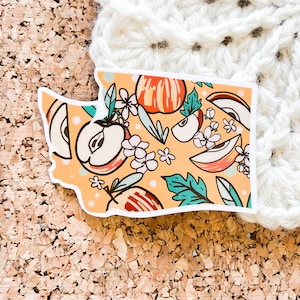 May include: A sticker featuring a stylized outline of the state of Washington, filled with a pattern of apples, flowers, and leaves in shades of red, orange, yellow, green, and white on a light orange background.