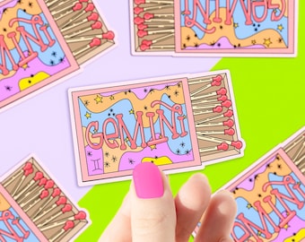 May include: A pink and blue matchbox sticker with the word "Gemini" and a stylized design of stars and swirls. The matchbox is open and shows a row of matches with red tips.
