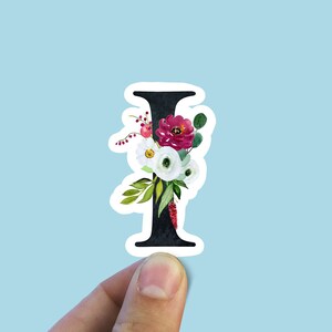 May include: A black letter I with a white outline, decorated with watercolor flowers in shades of pink, white, and green. The flowers are arranged around the letter, creating a floral bouquet.