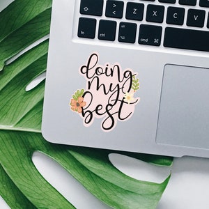 May include: A pink sticker with the text "doing my best" in black cursive lettering. The sticker is decorated with small orange and yellow flowers and green leaves.
