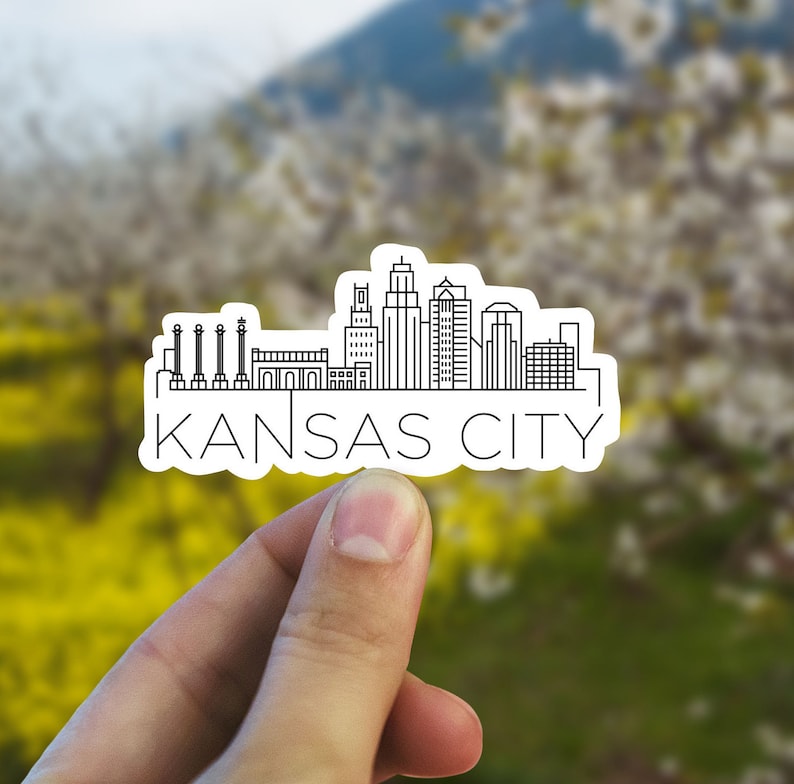 K&ouml;nnte beinhalten: Ein wei&szlig;er Aufkleber mit einer schwarzen Strichzeichnung der Skyline von Kansas City. Das Wort "KANSAS CITY" ist darunter in Schwarz gedruckt. Der Aufkleber wird in einer Hand vor einem verschwommenen gr&uuml;n-gelben Hintergrund gehalten.