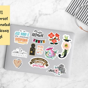 May include: A laptop with a variety of stickers on it. The stickers feature different designs, including flowers, text, and illustrations. The text on the stickers includes "Just Wing It", "I'm Best Served With Coffee and a Side of Sarcasm", "My Fountain Alcohol", and "Stockholm".