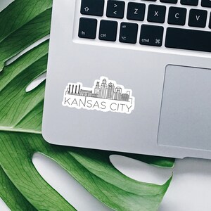 K&ouml;nnte beinhalten: Ein wei&szlig;er Aufkleber mit einer schwarzen Strichzeichnung der Skyline von Kansas City, einschlie&szlig;lich Geb&auml;uden und Schornsteinen. Die Worte "KANSAS CITY" sind darunter gedruckt. Der Aufkleber befindet sich auf einem silbernen Laptop.