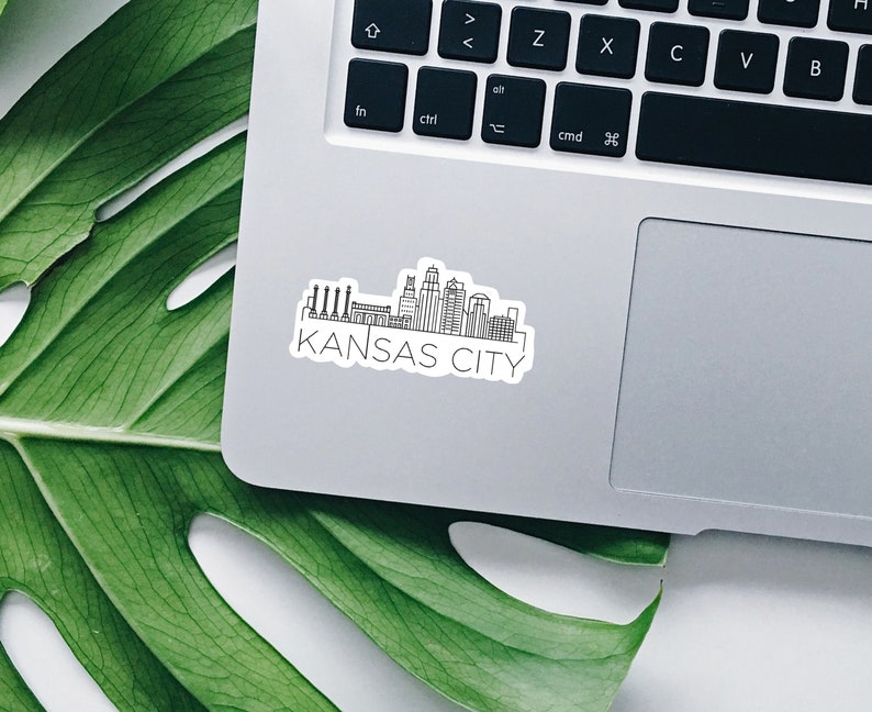 K&ouml;nnte beinhalten: Ein wei&szlig;er Aufkleber mit einer schwarzen Strichzeichnung der Skyline von Kansas City, einschlie&szlig;lich Geb&auml;uden und Schornsteinen. Die Worte "KANSAS CITY" sind darunter gedruckt. Der Aufkleber befindet sich auf einem silbernen Laptop.