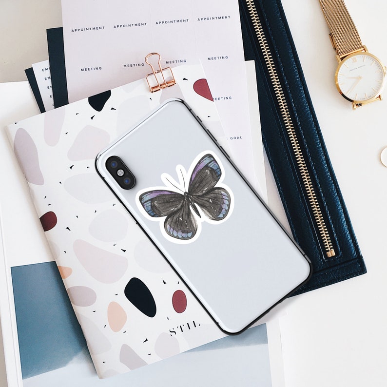 Op de afbeelding: Een smartphone met een vlindersticker, liggend op een notitieboek met een terrazzo patroon. De vlindersticker is zwart met paarse en blauwe accenten. Een gouden horloge en een donkerblauwe portemonnee zijn ook zichtbaar.