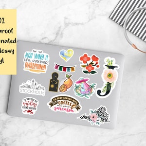 K&ouml;nnte beinhalten: Eine Sammlung wasserfester, UV-laminierter, dick gl&auml;nzender Vinyl-Aufkleber auf einem Laptop. Die Aufkleber zeigen verschiedene Designs, darunter florale Motive, Zitate und Illustrationen. Ein Kopfh&ouml;rer ist ebenfalls zu sehen.