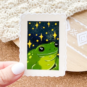 May include: A green frog with black eyes and white spots is looking up at a starry night sky. The frog is surrounded by yellow stars and a dark blue background. The image is framed with a white border.