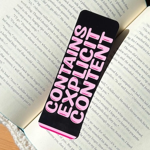 May include: Black bookmark with the text "CONTAINS EXPLICIT CONTENT" in pink, placed on an open book. The bookmark is rectangular and has rounded corners. The book's pages are yellowed.