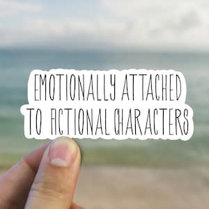 May include: A white sticker with the text "EMOTIONALLY ATTACHED TO FICTIONAL CHARACTERS" in black, held against a blurred ocean backdrop. The sticker has a rounded rectangular shape.