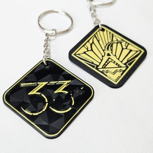 May include: Two black keyrings with silver chains. One keyring features the number "33" in yellow, the other has a yellow design. Both keyrings are diamond-shaped with a black background and yellow trim.