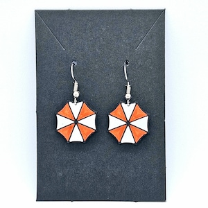 May include: A pair of silver earrings with white and orange umbrella-shaped charms. The charms feature a black outline and a white background with an orange, eight-pointed star.