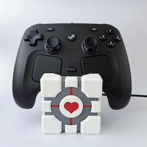 以下が含まれることがあります： 黒いSteamコントローラーが、ハートのシンボルが付いた白、グレー、赤のキューブの後ろに配置されています。コントローラーには2つのジョイスティック、方向パッド、いくつかのボタンがあります。キューブの中央にはハートがあります。
