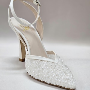 May include: White high-heeled shoe with a pointed toe and ankle strap. The toe box is covered in small, white, pearl-like beads. The heel is a thick, square shape. The shoe has a buckle closure on the ankle strap.