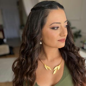 May include: A gold leaf necklace with a fine chain. The necklace is worn over a green top. The woman has long, wavy brown hair and is wearing makeup. A pearl earring is visible.
